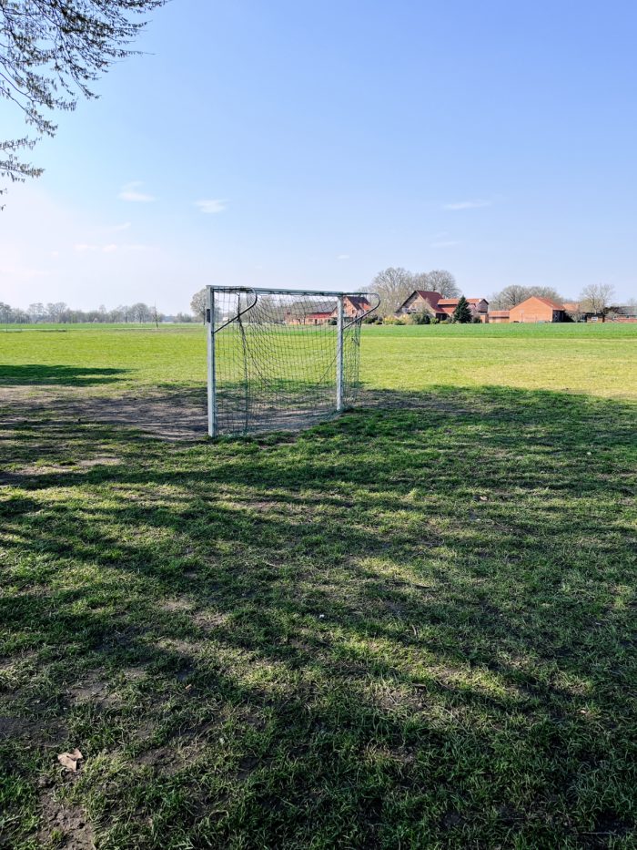 Ein Fußballtor steht auf einer weiten, leeren Rasenfläche, im Hintergrund ziegelrote Bauernhäuser