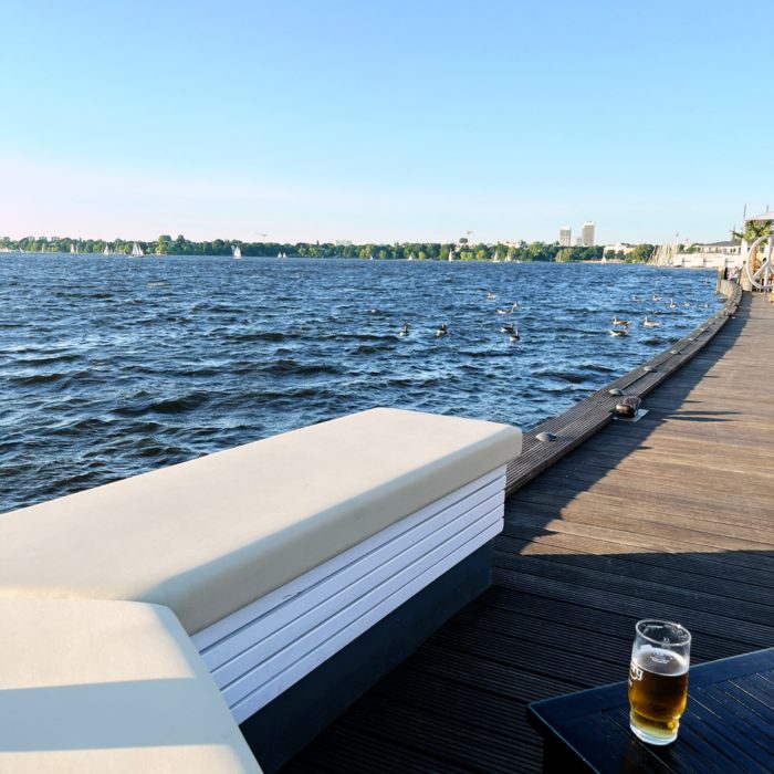 Ein Steg an der Alster, eine Bank mit Sitzpolster darauf, daneben ein Glas Bier