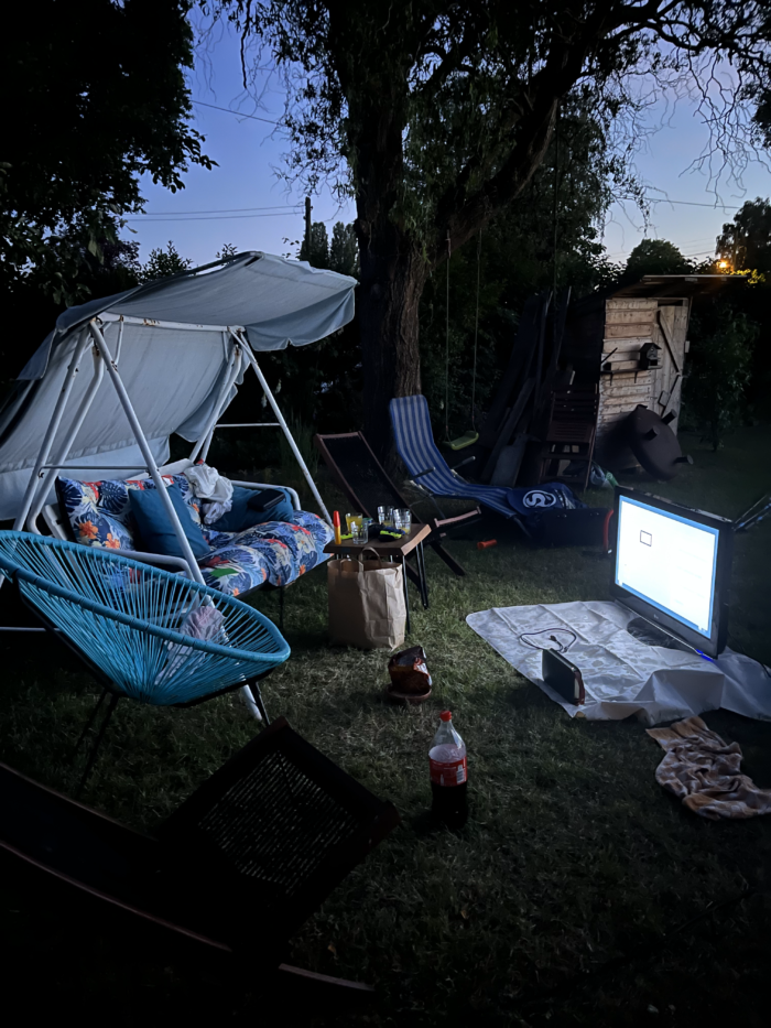 Eine Hollywoodschaukel im dunklen Garten, davor ein großer Monitor und Gaming-Zubehör