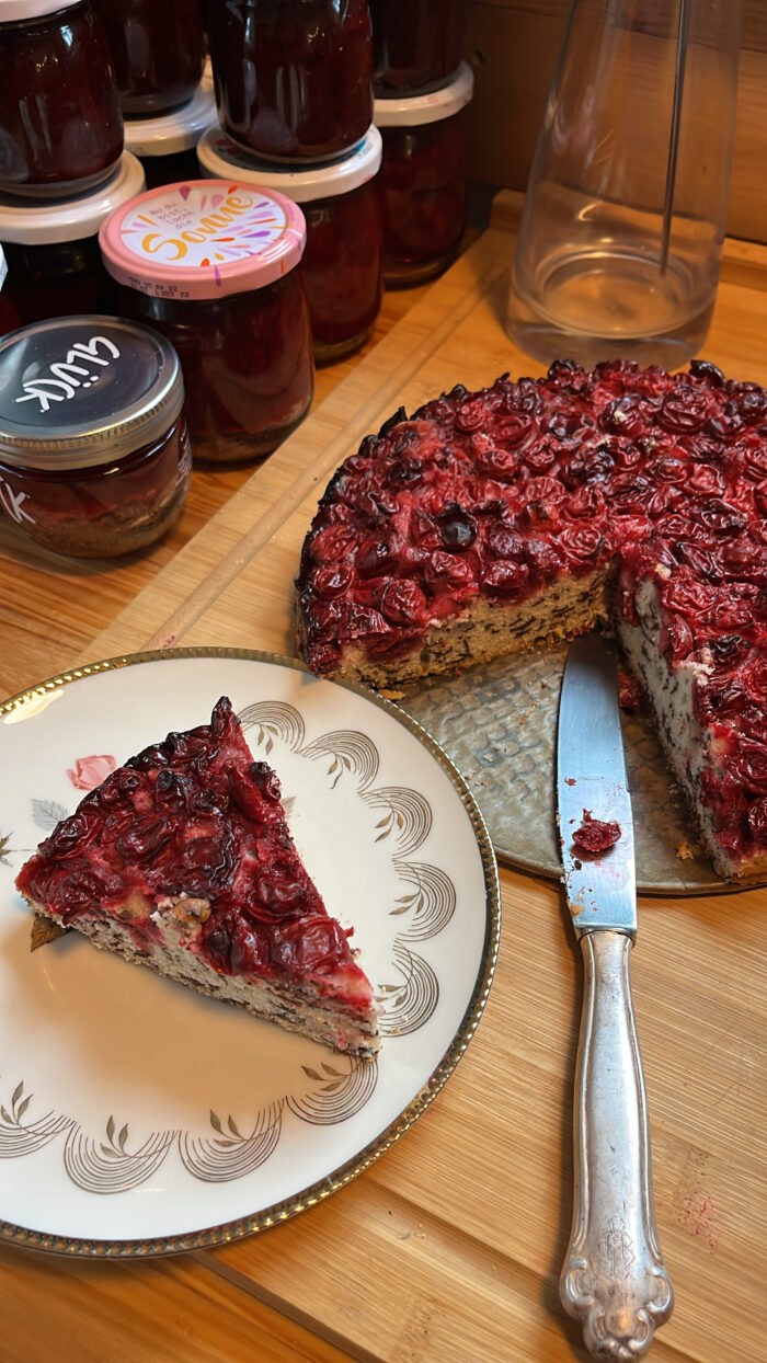 Ein Kirschkuchen, ein Stück auf einem Teller, daneben Gläser mit frischer Kirschmarmelade
