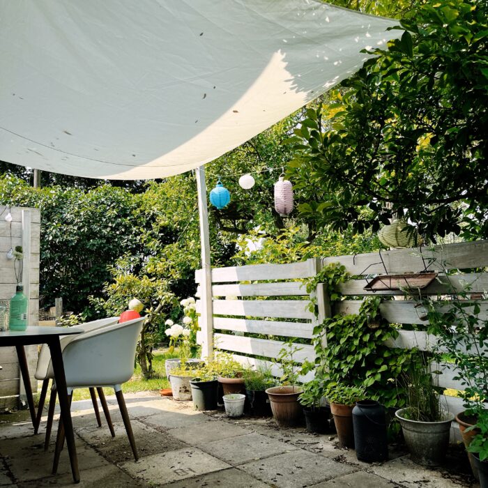 Eine Gartenszene, eine Terrasse, Tisch, Stuhl, Lampions, ein Sonnensegel darüber, ein sonniger Tag