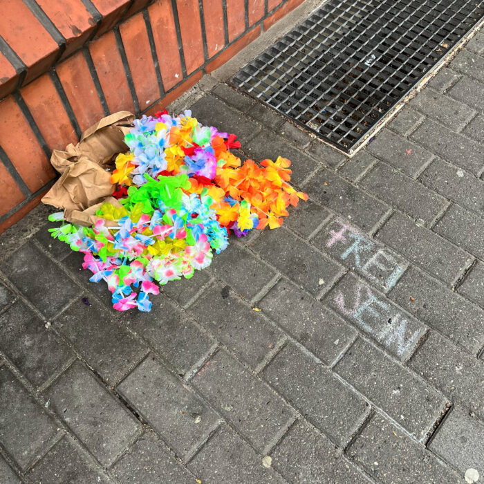 Bunte Kreideschrift auf einem Gehweg: "Frei Sein", daneben eine Plastikblumengirlande