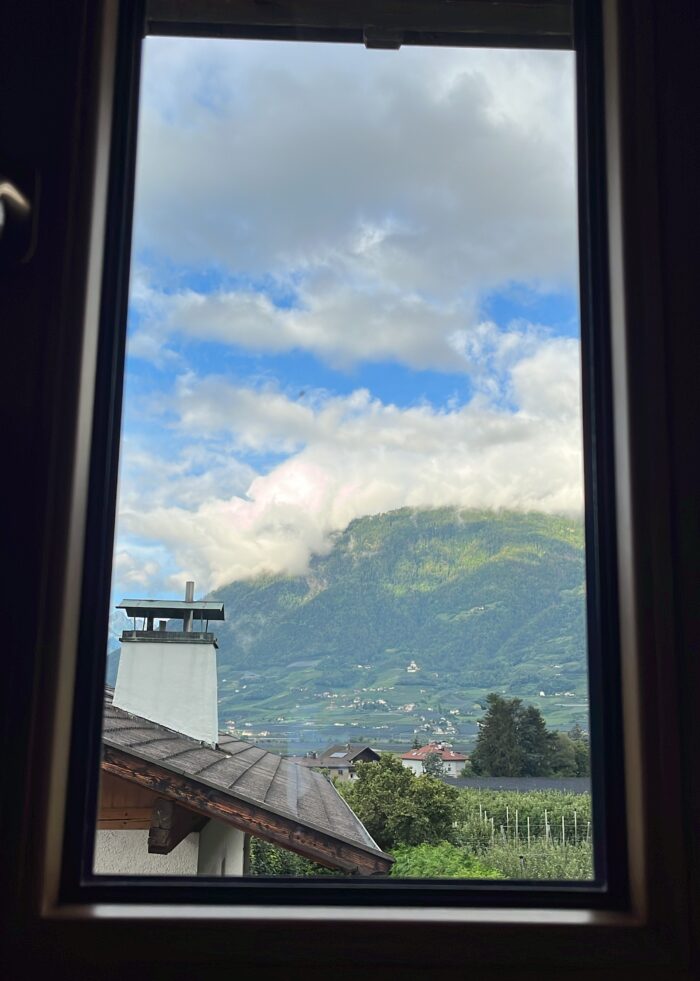 Blick durch ein schmales Fenster über ein Dach mit Schornstein auf Berge im Hintergrund, bewölkter Himmel, Weinstöcke in der Landschaft