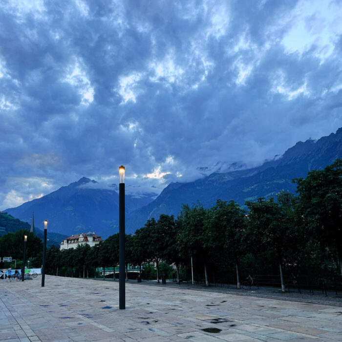 Abendlicht an der Promenade an Meran, eine erleichtete Laterne vor den Bergen im Hintergrund der Stadt, blaue Stunde