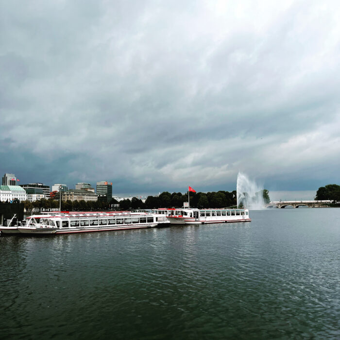 Zwei Schiffe der weißen Flotte auf der Binnenalster vor herbstlich graudunklem Himmel, im Hintergrund das Finnlandhaus und die Alsterfontäne, ein ICE auf der Brücke fährt gerade zum Hauptbahnhof, eine rot leuchtende Hamburgflagge