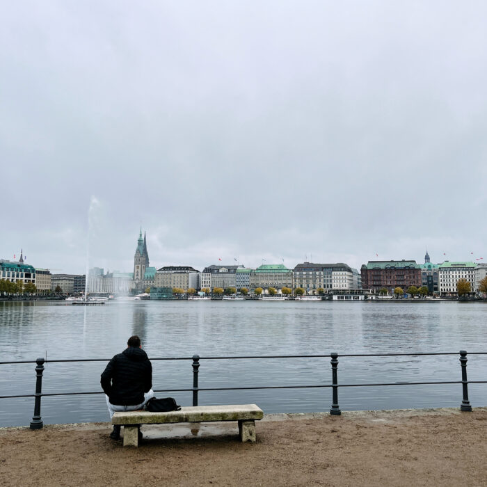 Blick über die Binnenalster Richtung Jungfernstieg, Rathaus. Im Vordergrund ein Mann auf einer Bank, man sieht ihn von hinten.