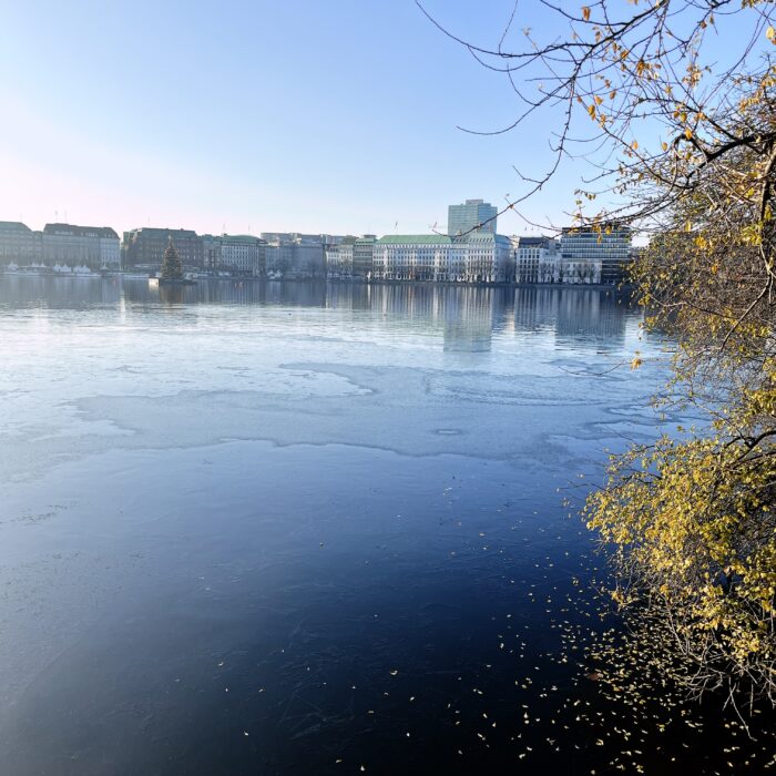 Blick über die Binnenalster auf den Jungfernstieg, dünnes, frisches Eis im Vordergrund, es reicht nicht über die ganze Fläche