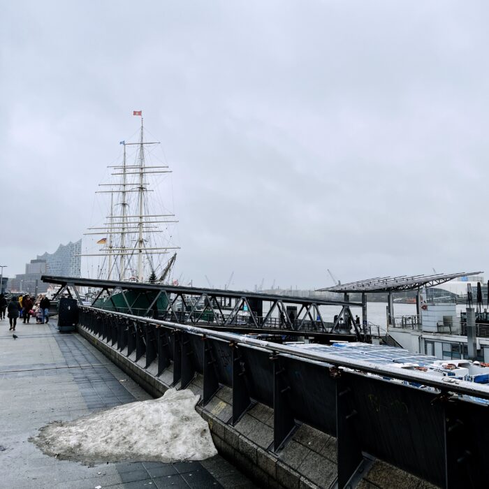 Ein schmuddeliger Schneerest auf der Hafenpromenade, kurz vor der Rickmer Rickmers, im Hintergrund die Elbphilharmonie vor grauem Himmel