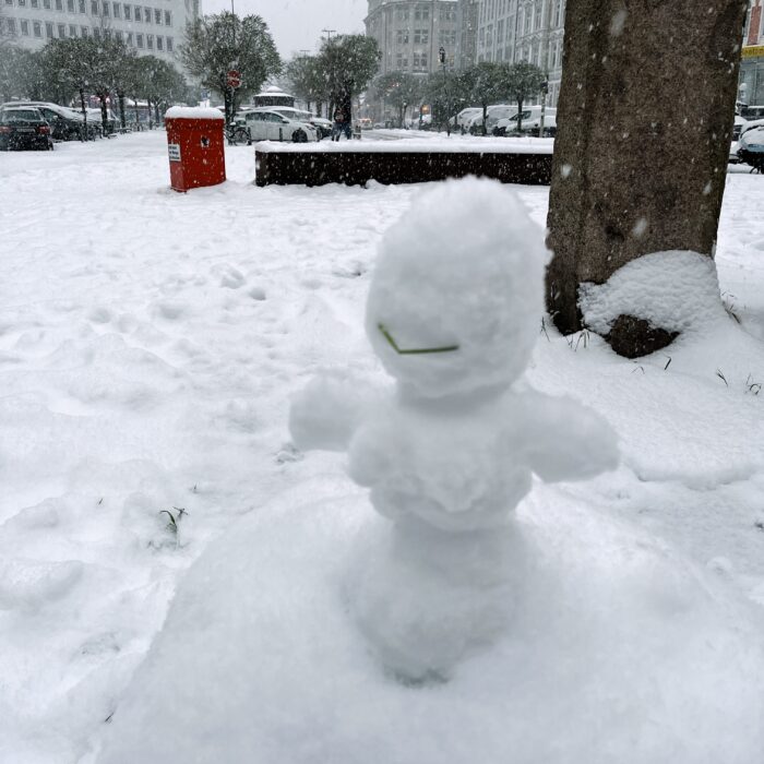 Eine kleine Schneefigur mit deutlichen Brüsten und einem geknickten Grashalm als Lächeln im Gesicht