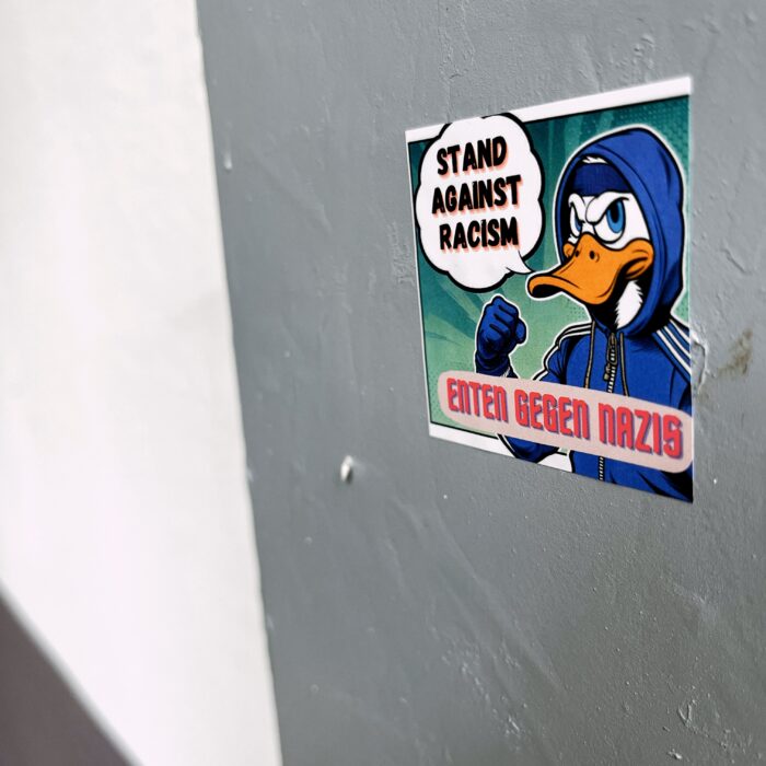 Ein Sticker an einer Wand, darauf eine Ente im Disney-Stil, wie Donald, mit Sprechblase: "Stand against Racism" , darunter der Slogan "Enten gegen Nazis"