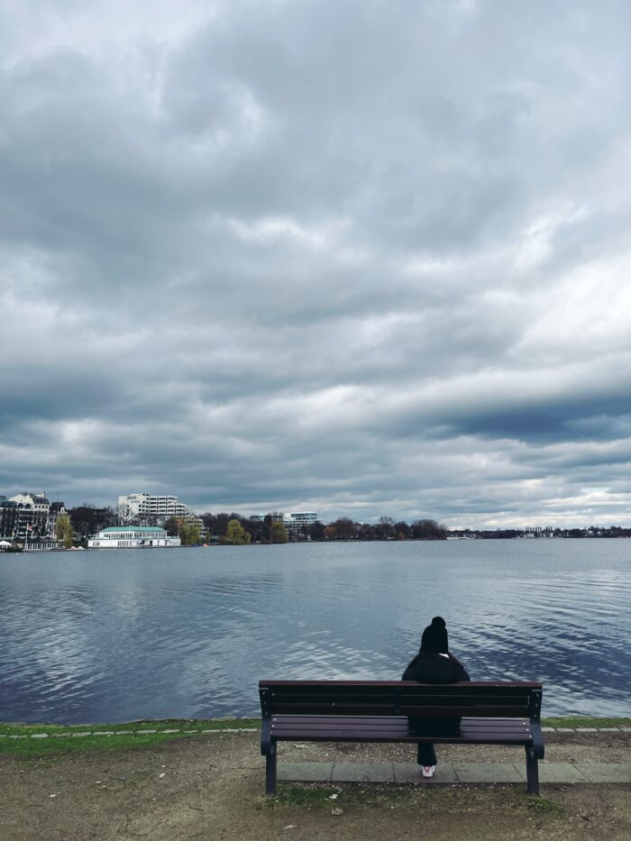 Jemand sitzt auf einer Bank an der Außenalster, die Person wurde von hinten aufgenommen. Viele Wolken am Himmel, aber die gebäude am Ufer erscheinen hell und wie angestrahlt, es kommt Licht durch.