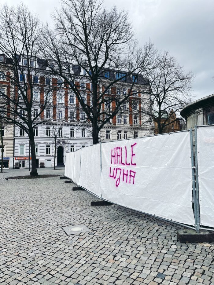 Ein mit weißer Folie bespannter Bauzaun, auf den jemand in pinkfarbener Schrift "Hallelujha" geschrieben hat, mit Rechtschfreibfehler und so getrennt, dass oben Halle und unten Lujha steht. Der Bauzaun steht auf einem sonst leeren Platz, der Schriftzug wirkt dadurch sehr prominent.