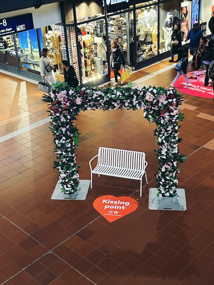 Eine weiße Bank in der Wandelhalle, ein Plastikblumenbogen darüber, davor steht auf dem Boden in einem Herz: Kissing Point