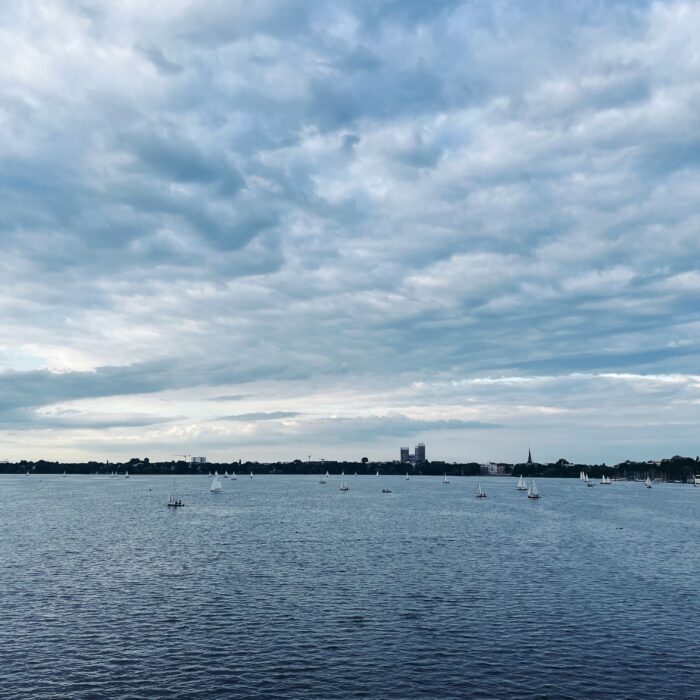 Blick von der Kennedybrücke auf die Außenalster unter blaugrau verhangenem Himmel. Einige Segelboote, im Hintergrund die Mundsburghochhäuser.