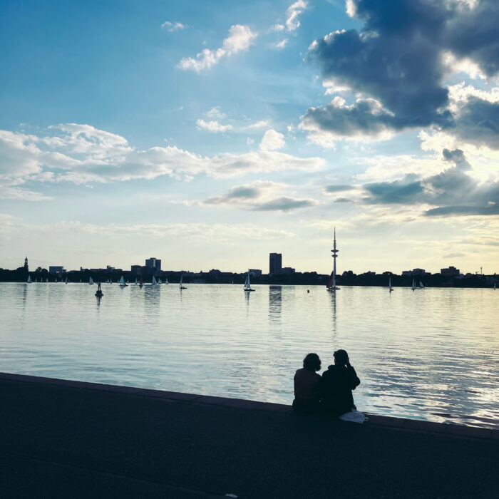 Ein Pärchen sitzt im letzten Abendlicht am Ufer der Außenalster und blickt übers Wasser. Die beiden sind von hinten nur als Silhouetten zu sehen. Am Horizont, am anderen Ufer der Fernsehturm.