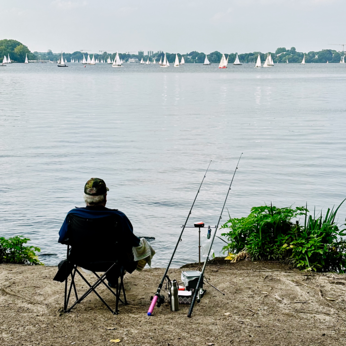 Blick auf die Außenalster von der St-Georg-Seite aus. im Vordergrund ein Angler am Ufer, er sitzt in einem Klappstuhl, bene sich zwei aufgestellte Angeln. Im Hintergrund etliche weiße Segel.