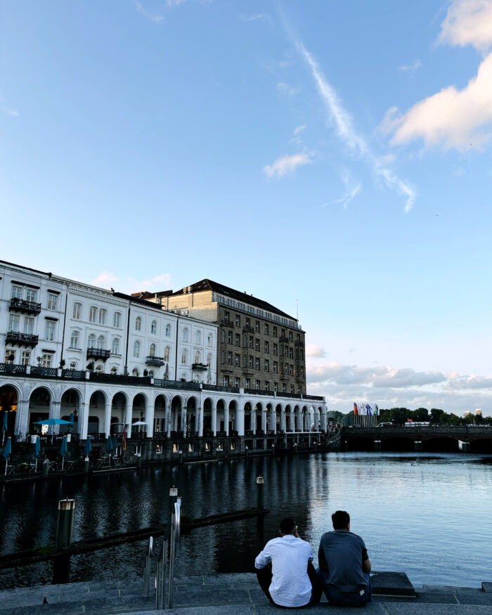 Die Hamburger Alsterarkaden und die Kleine Alster. Zwei Menschen sitzen auf den Treppen am Ufer, man sieht sie von hinten. Sommerliche Anmutung, blauer Himmel, Dekowölkchen.