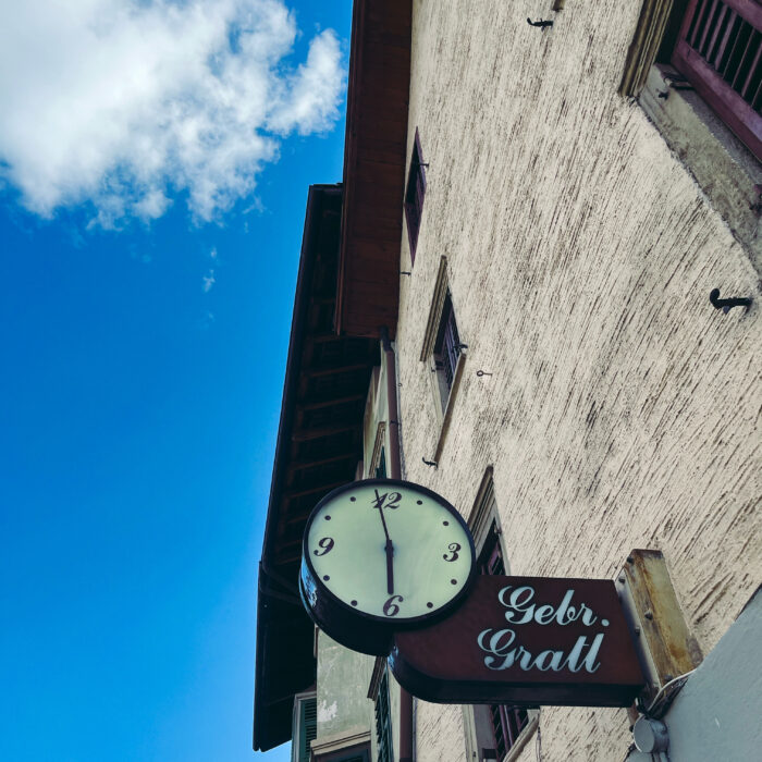 Eine altmodische Uhr an einem Juwliergeschäft in Kaltern, darunter der Schriftzug "Gebr. Grattl" in ebenfalls alter Typo, geschwungene Schreibschrift