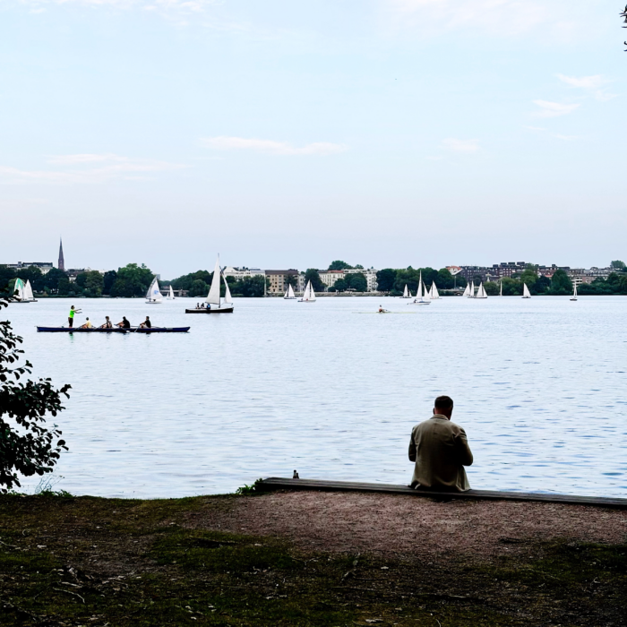 Ein Mann sitzt am Ufer der Ufenalster, Sankt-Georg-Seite, er ist von hinten zu sehen. Vor ihm Ruder- und Segelboote, Sommreabend.