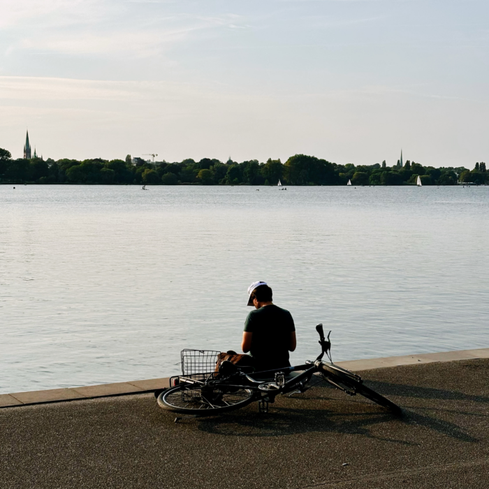 Ein Mann sitzt am Ufer der Außenalster, Höhe Schwanenwik, er hat sein Fahrrad hinter sich abgelegt und liest etwas.