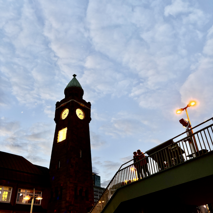 Der Uhrturm an den Landungsbrücken im Abendlicht, Passanten am Geländer der S-Bahnstation im Gegenlicht