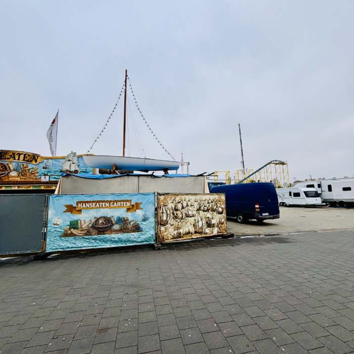 Ein Detail aus der etwas trostlos wirkenen Szenerie des Dom-Aufbaus, an einem Zaun ein buntes Plakat mit der Aufschrift "Hanseatengarten", darüber sieht man ein Segelboot, das irgendwo zu dekorativen Zwecken verarbeitet werden wird, daneben Parkplatzleere