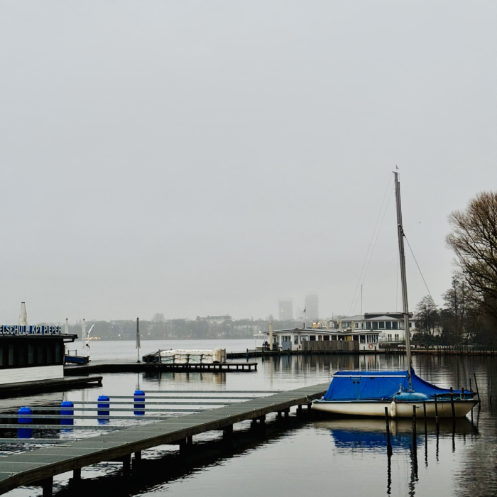 Die Außenalster vom Ufer St. Georg aus, ein mit Plane bedecktes Segelboot im Vordergrund, graues Nachmittagslicht, Dunst über der Stadt