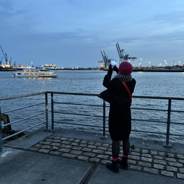 Die Herzdame, von hinten fotografiert, wie sie am Fischmarkt steht und über die Elbe mit dem Smartphone fotografiert. Später Nachmittag, es dunkelt schon, beleuchtete Fähren auf der Elbe, Hafenkräne im Hintergrund.