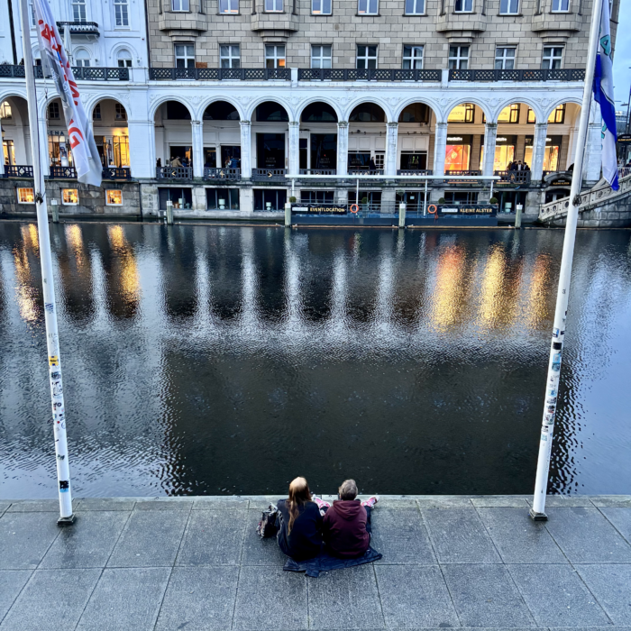 Ein Paar sitzt auf dem gepflasterten Ufer der Kleinen Alster und sieht auf die Alterarkaden