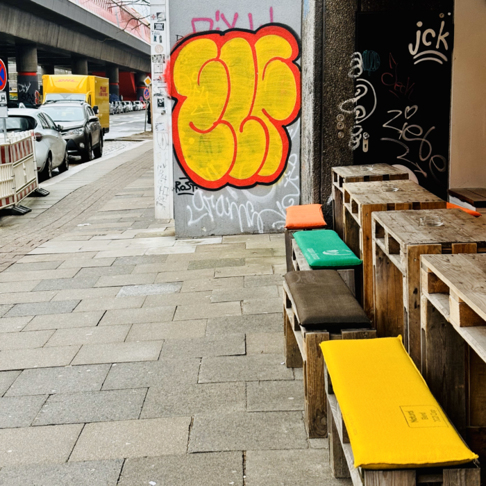 Eine Straße in Hammerbrook mit leuchten gelbrotem Graffiti-Tag und bunten Kissen auf den Bänken vor einem Imbiss