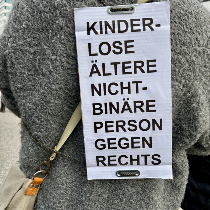 Ein Pappschild auf dem Rücken einer demonstrierenden Person: "Kinderlose ältere nicht -binäre Person gegen rechts"