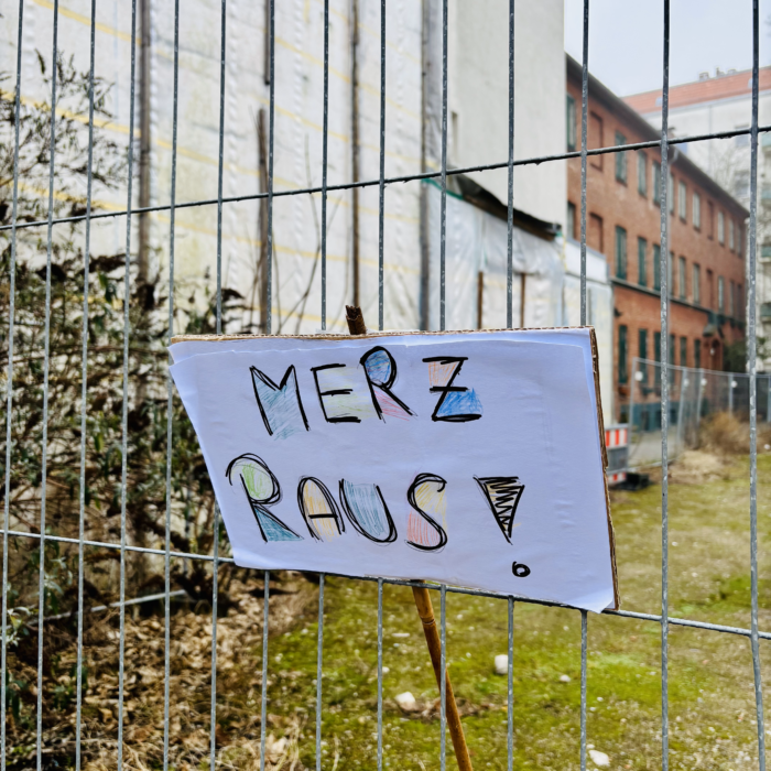 Ein Demoplakat mit dem Text "Merz raus" (Filzstifte, bunt), das in einen Bauzaun vor einem leeren Grundstück eingehängt wurde
