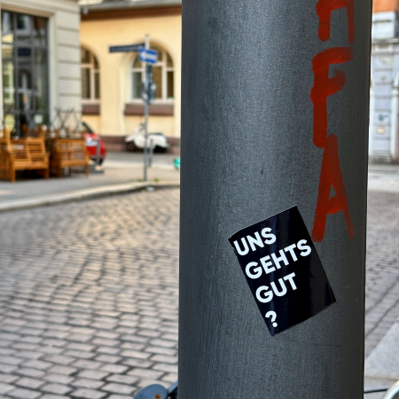 Ein Aufkleber mit dem Text "Uns gehts gut?" an einem Laternenmast