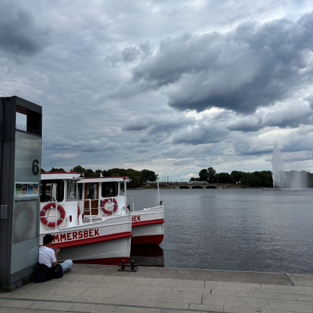 Blick vom Anleger Jungfernstieg auf die Binnenalster unter Regenwolken, ein Mann sitzt an der Kante und sieht Richtung Fontäne, zwei Alsterschifffe angeschnitten im Bild