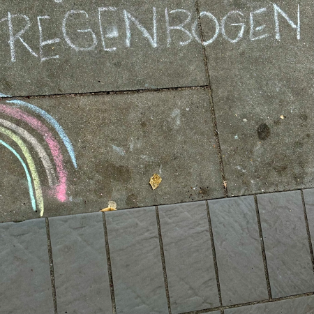 Eine Kinderkreidezeichnung auf dem Pflaster von einem Regenbogen, darüber das Wort Regenbogen in Großbuchstaben