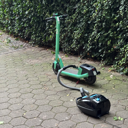 Ein Staubsauger steht neben einem E-Roller, es sieht aus, als seien sie durch den Schlauch des Staubsaugers in eindeutiger Weise verbunden