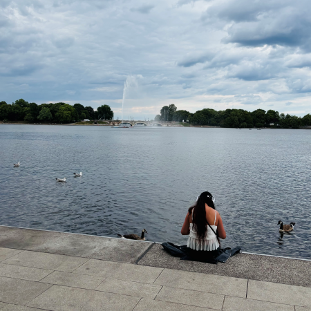 Eine Frau, von hinten gesehen, sitzt an der Binnenalster. Im Hintergrund die Fontäne.