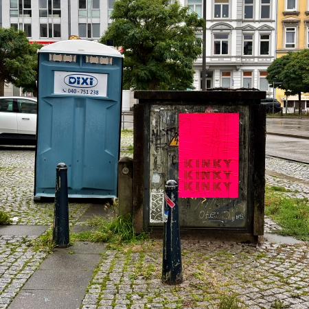 Ein Dixie-Klo neben einem Stromkasten, auf dem ein pinkfarbenes Plakat klebt, auf dem Kinky Kinky Kinky steht