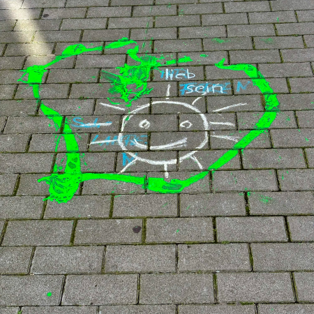Farbe und Kreide auf dem Pflaster, eine gemalte Sonne und der Text: Lieb sein.