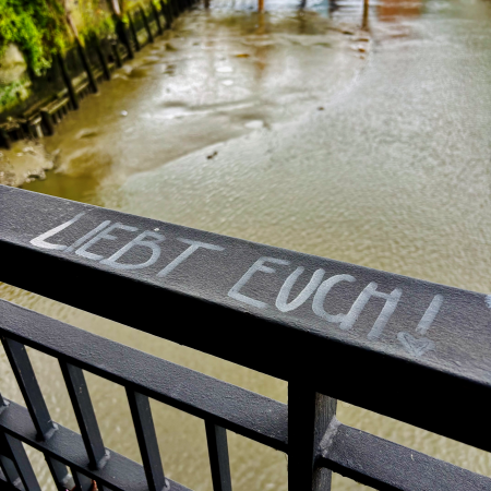 Weiße Schrift auf einem eisernen Brückengeländer: "Liebt Euch!"