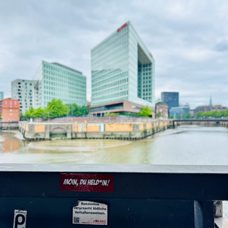 Blick auf das Hamburger Spiegelgebäude, auf einem Brückengeländer im Vordergrund klebt ein Sticker mit Text: "Moin, Du Held*in!