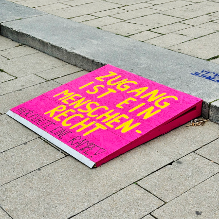 Eine improvisierte Rampe für Rollstuhlfahrerinnen an einer Bordsteinkante mit dem Text "Zugang ist ein Menschenrecht, hier fehlte eine Rampe"