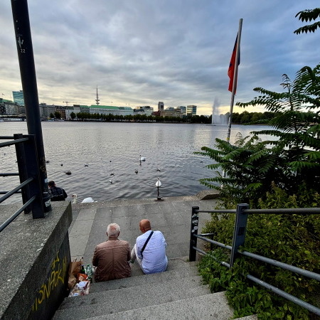 Zwei Männer, von hinten gesehen, die auf den Stufen sitzen, die am Ballindamm hinunter zur Binnenalster führen, im Hintergrund Fontäne und Fernsehturm