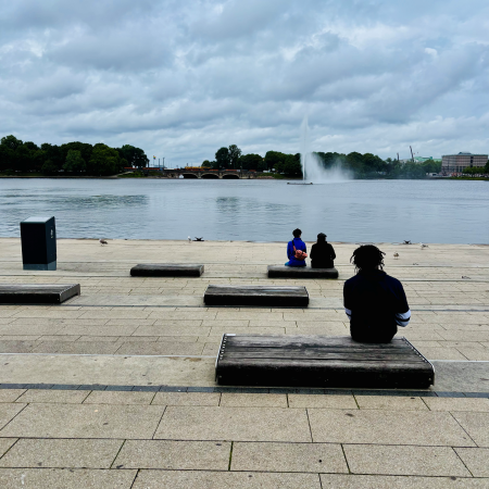 Drei Menschen, von hinten gesehen, sitzen auf den Bänken vor der Binnenalster am Jungfernstieg, im Hintergrund die Fontäne
