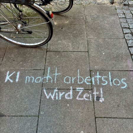 Kreideschrift auf dem Pflaster: "KI macht arbeitslos - wird Zeit!"