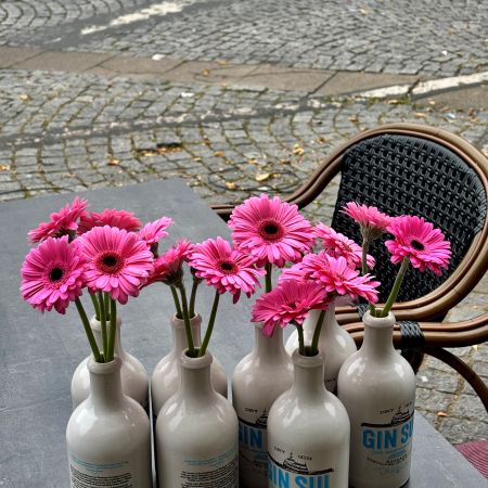 Etliche pinkfarbene Gerbera in Ginflaschen als Vasen auf einem Tisch in der Außengastro
