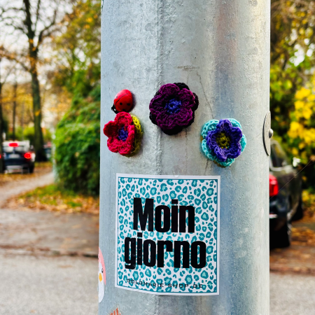 Ein Aufkleber an einem Laternenpfahl "Moin Giorno", darüber angeklebte Strickblüten