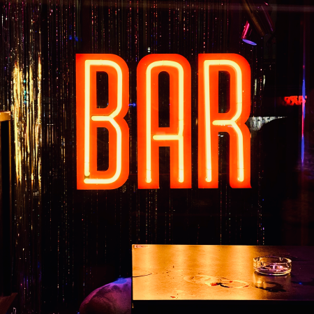 Ein nachts leuchtendes Neonschild "BAR", davor ein Aschenbecher auf einem Tisch vor der Kneipentür