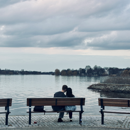 Ein Paar auf einer Bank an der Außenalster, von hinten fotografiert, sie halten Kopf an Kopf