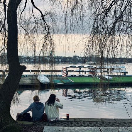 Ein Paar mit Getränken am Ufer der Außenalster, von hinten fotografiert, vor ihnen Bootsstege